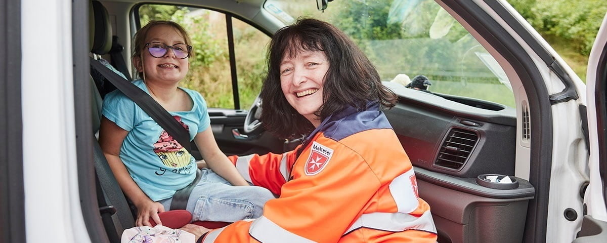 Eine Mitarbeiterin aus dem Fahrdienst grinst mit dem Mädchen, welches im Auto sitzt, zusammen in die Kamera.