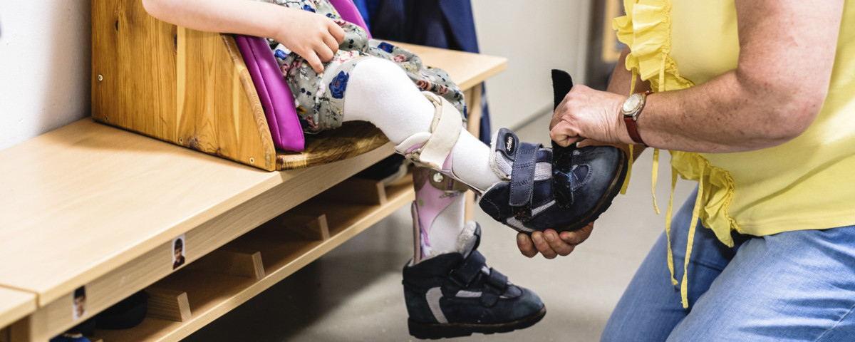 Belgeiterin/ Erzieherin hilft einem Kind mit Behinderung beim anziehen der Schuhe. 